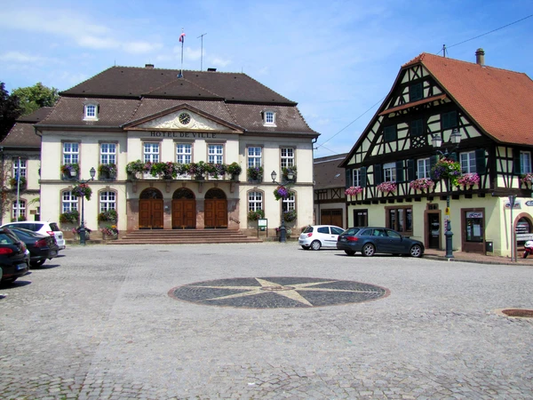 Vue aérienne ou paysage urbain d'Erstein montrant l'architecture typique alsacienne avec bâtiments résidentiels et commerciaux, toitures traditionnelles, rue principale ou place de la ville avec quelques monuments ou bâtiments caractéristiques. L'image doit capturer l'essence d'une ville du Bas-Rhin, sans personnages, avec une qualité photographique professionnelle.