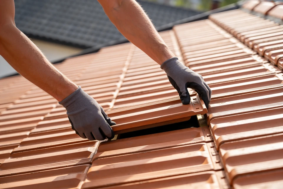 Vue d'une maison avec toiture en tuiles ou ardoise, légèrement endommagée ou nécessitant des travaux, prise de l'extérieur en lumière naturelle. Au premier plan, une personne en tenue professionnelle (artisan ou client) et un professionnel en équipement de sécurité discutent devant la maison, tenant des documents ou un devis. L'atmosphère doit transmettre la confiance, la facilité d'accès aux solutions de financement et la collaboration constructive entre le professionnel et son client.